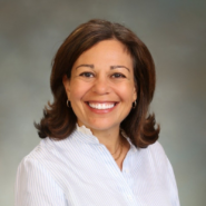 Headshot of a smiling person with shoulder-length hair, wearing a light-colored, collared shirt. The background is a soft gradient.