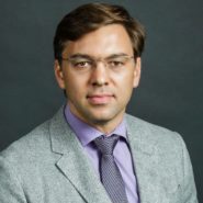 Headshot of a person with glasses, wearing a gray suit, lavender shirt, and patterned tie, set against a dark background.