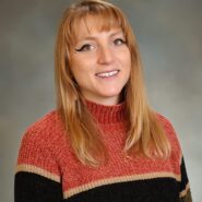 Headshot of a person with long hair, wearing a striped sweater in red, black, and beige. The background is plain with a neutral tone.