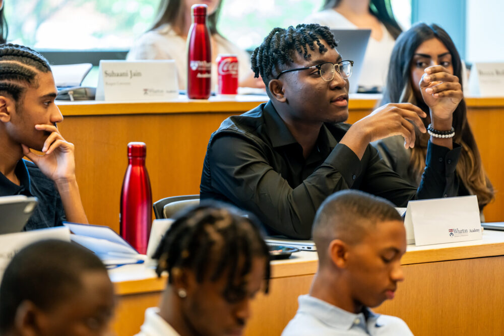 A group of students in a classroom setting, engaged in a discussion. One student is speaking while others listen attentively. Name placards and red water bottles are visible on the tables.