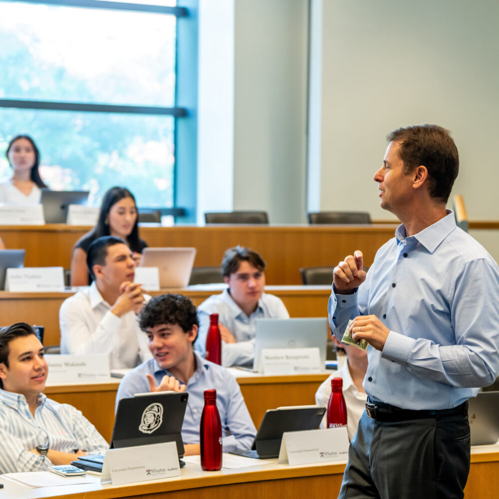 A classroom setting with a lecturer speaking to students sitting at desks with laptops and nameplates. The environment suggests a university lecture or seminar.