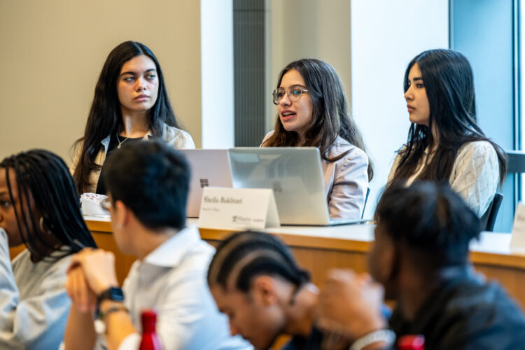 A group of young adults sitting in a classroom setting, with laptops and name cards in front of them. One person is speaking while others listen attentively.