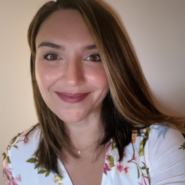 Headshot of a person smiling, with shoulder-length brown hair, wearing a floral-patterned top.