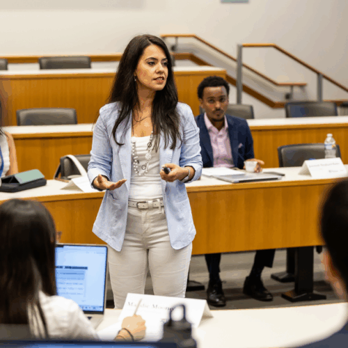A person standing and speaking in front of seated individuals in a classroom or seminar setting.
