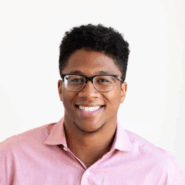Andre Douglas Headshot A person with short curly hair and glasses, smiling, wearing a pink shirt against a plain white background.